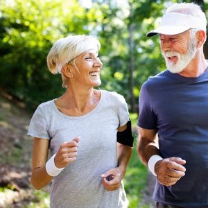 Elderly woman and man smile at each other while jogging on a sunny forest trail.